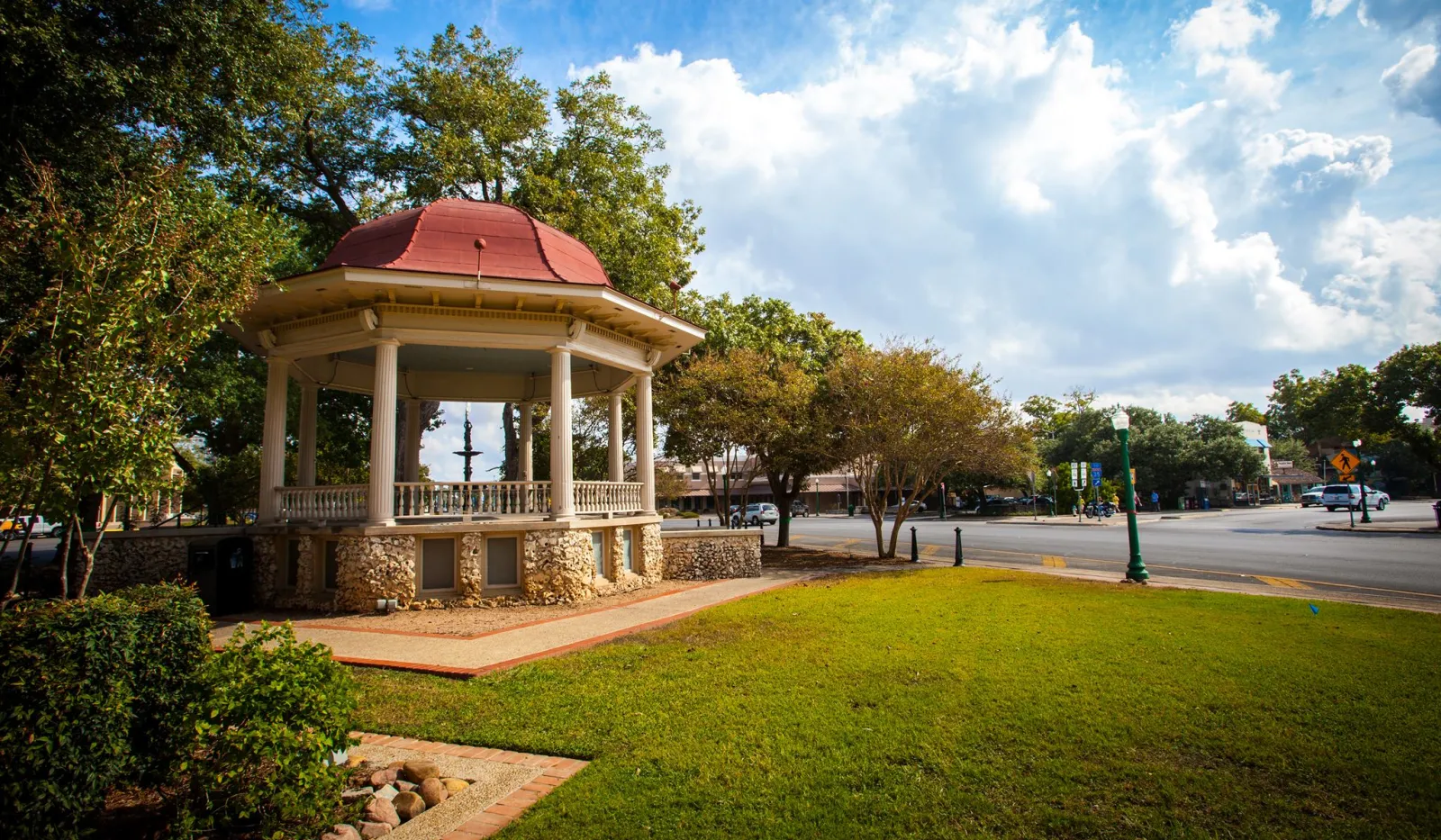 Octagonal gazebo with red roof in a park surrounded by green grass, trees, and a partly cloudy blue sky.