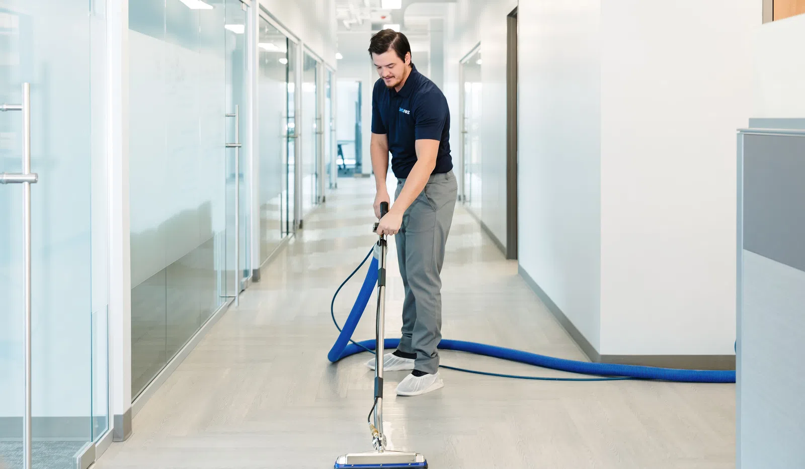 Man using professional carpet cleaning machine in a modern office corridor with glass walls and bright lighting.
