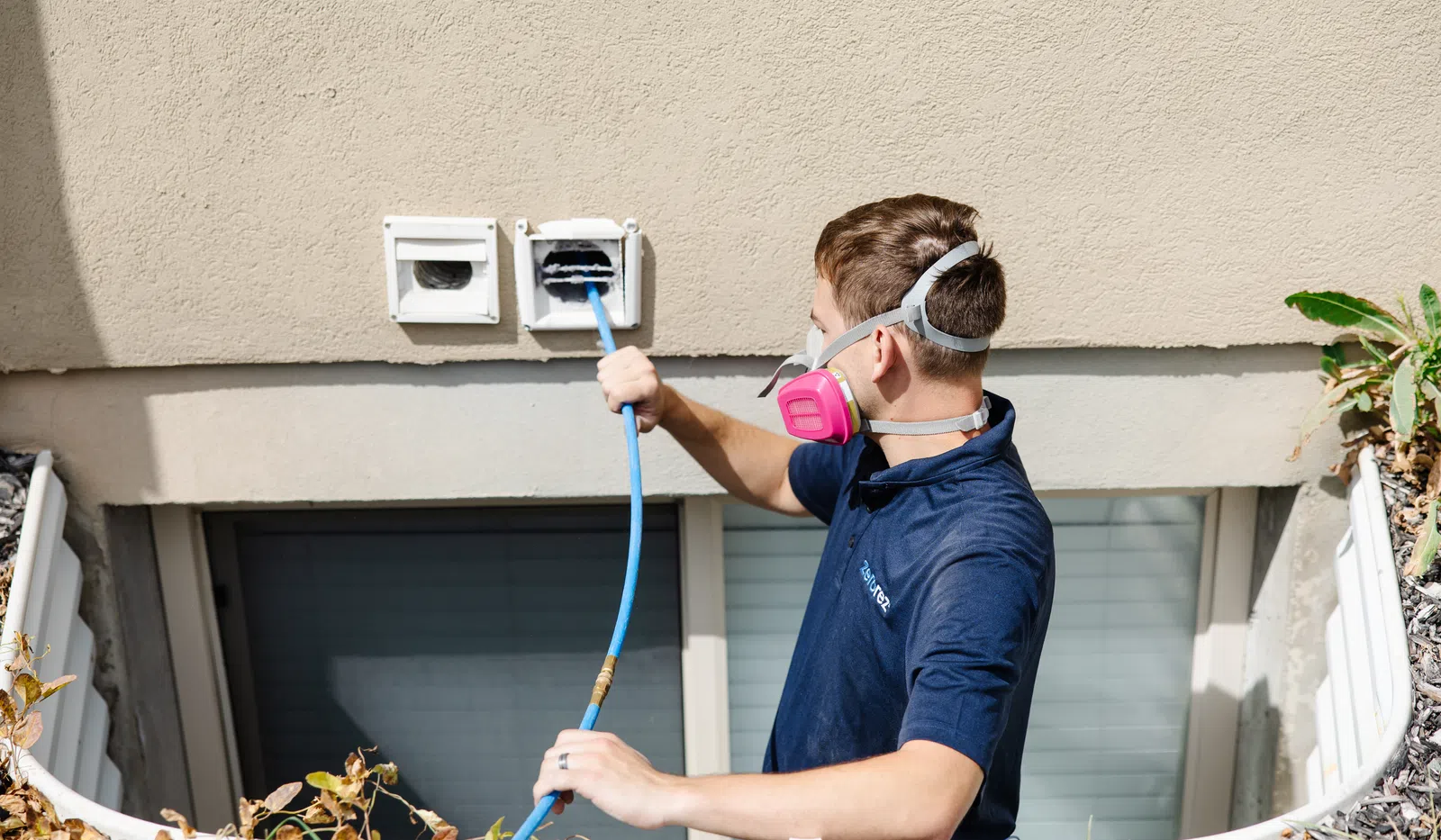 Technician in mask cleaning a dryer vent hose on a residential building exterior.
