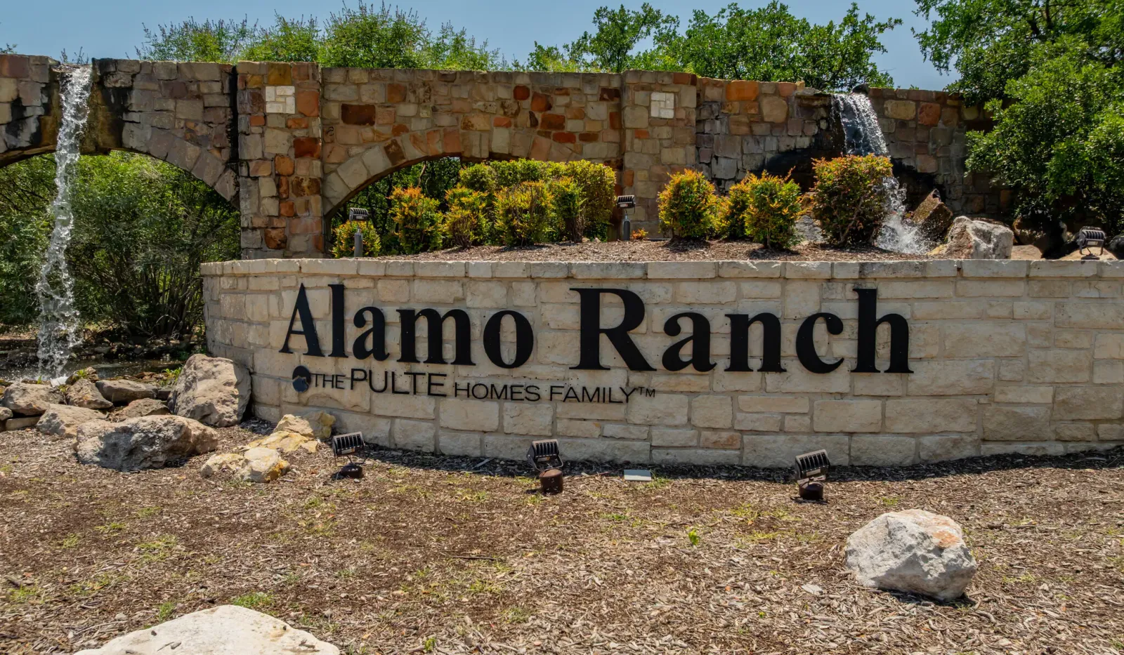 Stone entrance sign for Alamo Ranch with water features and greenery under a blue sky.