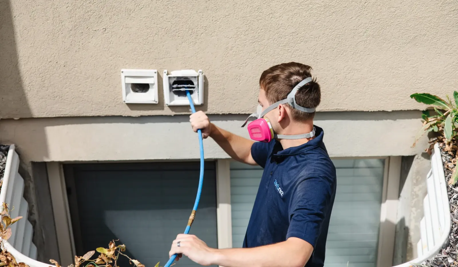 Technician wearing protective mask cleans dryer vent outside a house using a brush attached to a blue hose.
