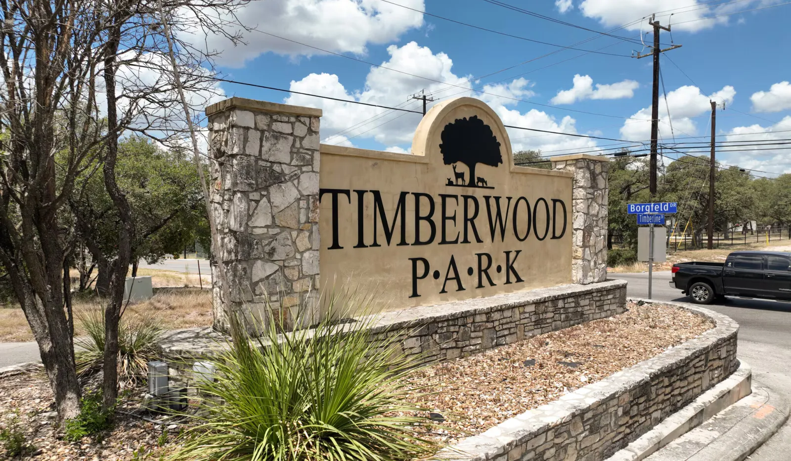 Stone and plaster entrance sign for Timberwood Park with blue sky and street in background.