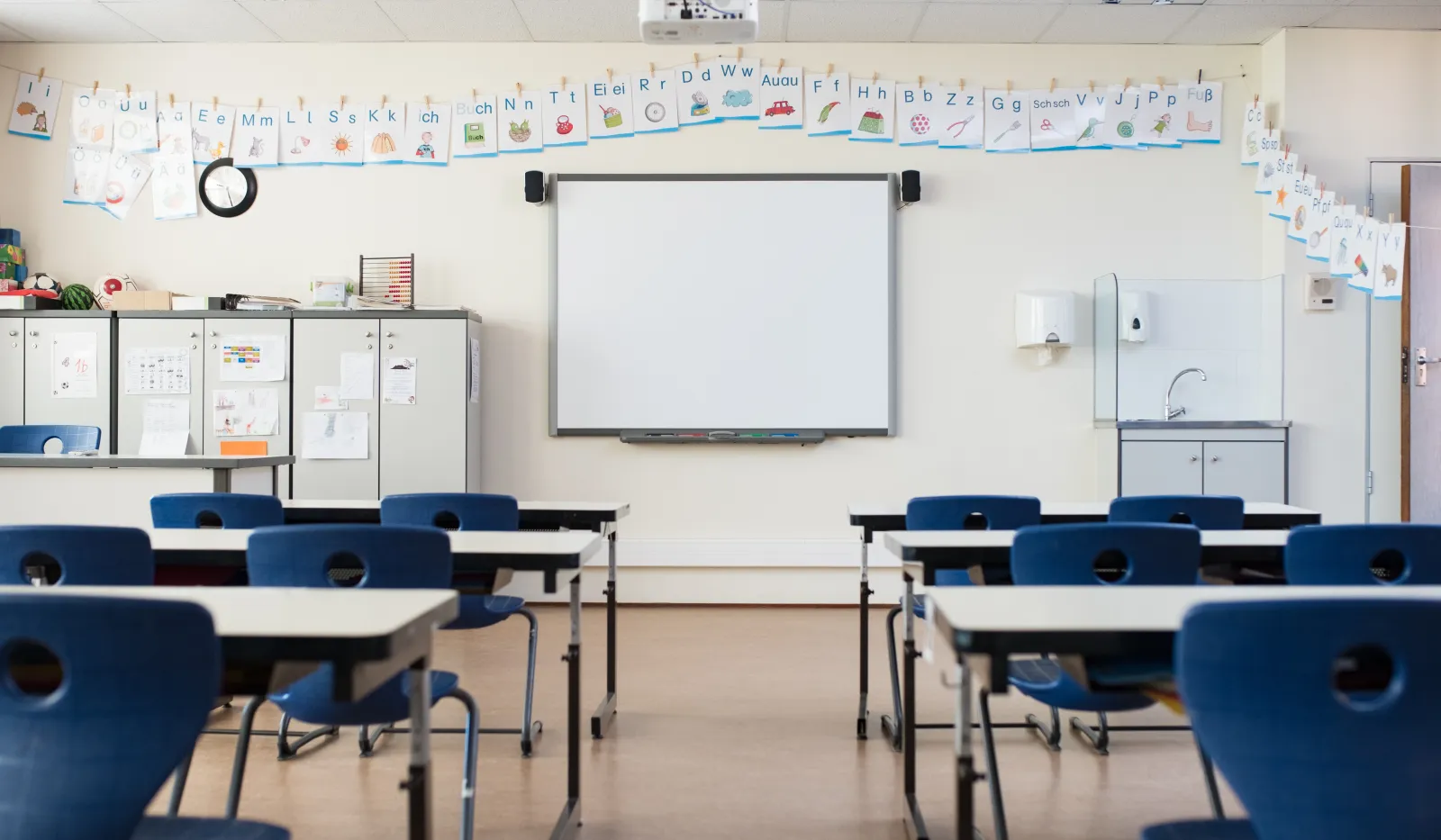 Empty classroom with blue chairs, desks, whiteboard, alphabet cards, and educational posters on walls.