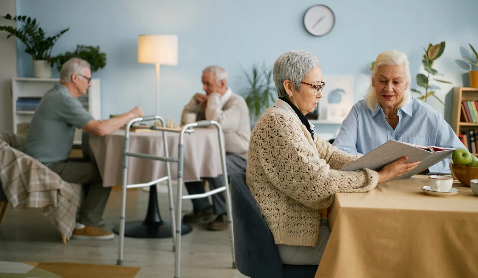Two elderly women reading a photo album at a table in a cozy living room with two men in the background.