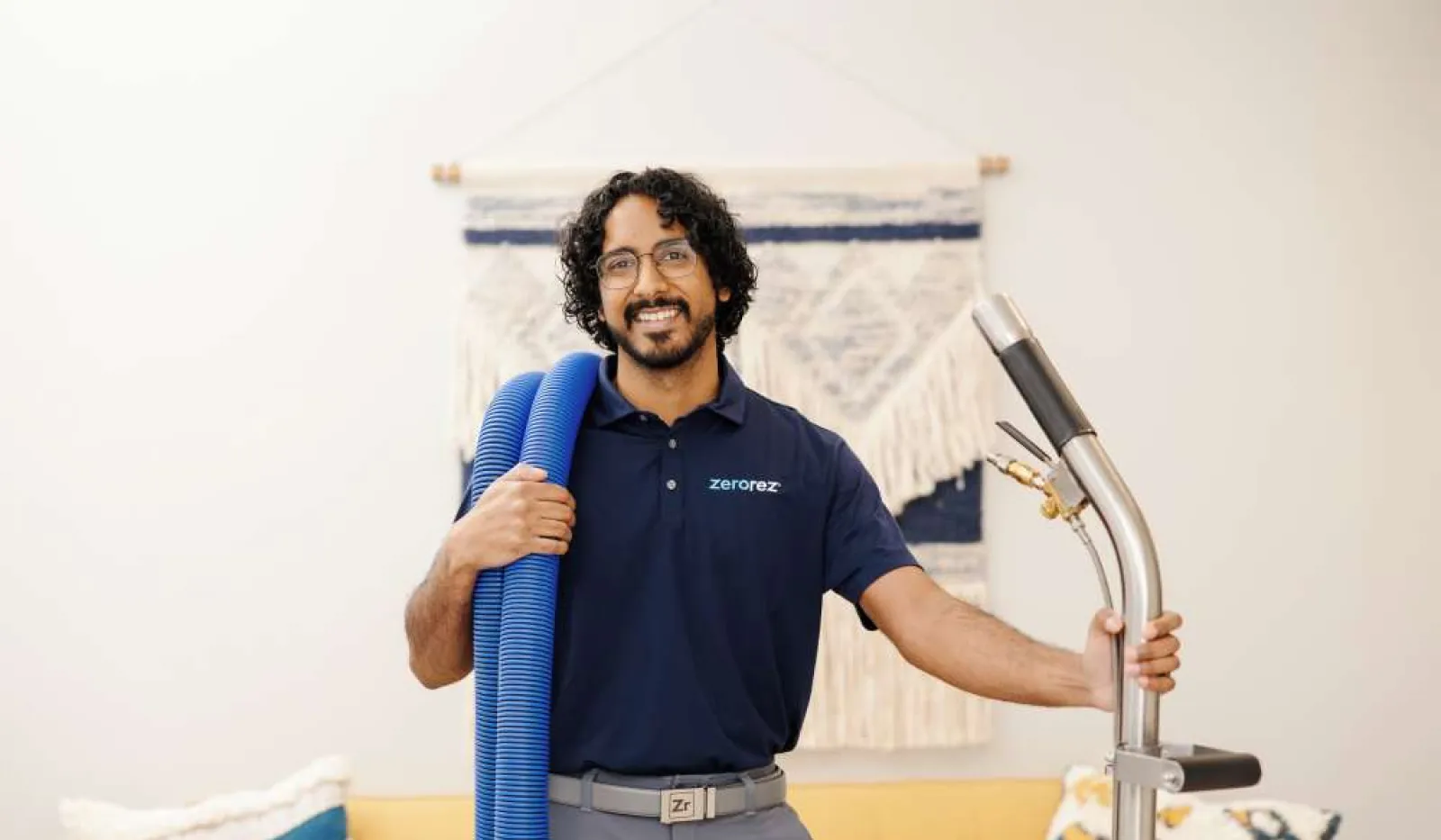 Smiling man in blue Zerorez shirt holding blue hose and cleaning equipment in a bright living room.