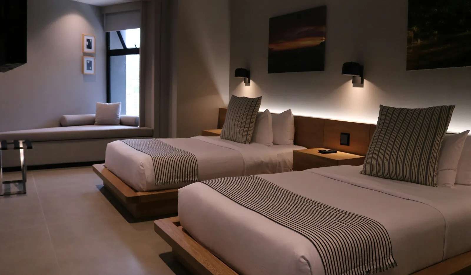 Modern hotel room with two beds featuring striped pillows and runners, ambient lighting, and a window seat.