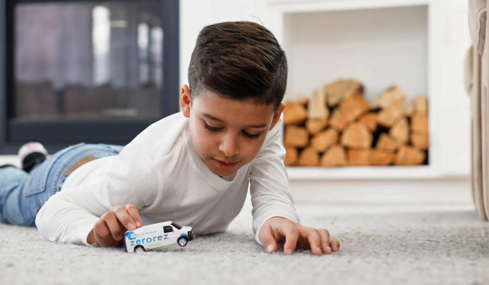 Young boy lying on carpet playing with a Zerorez branded toy van in a cozy living room.