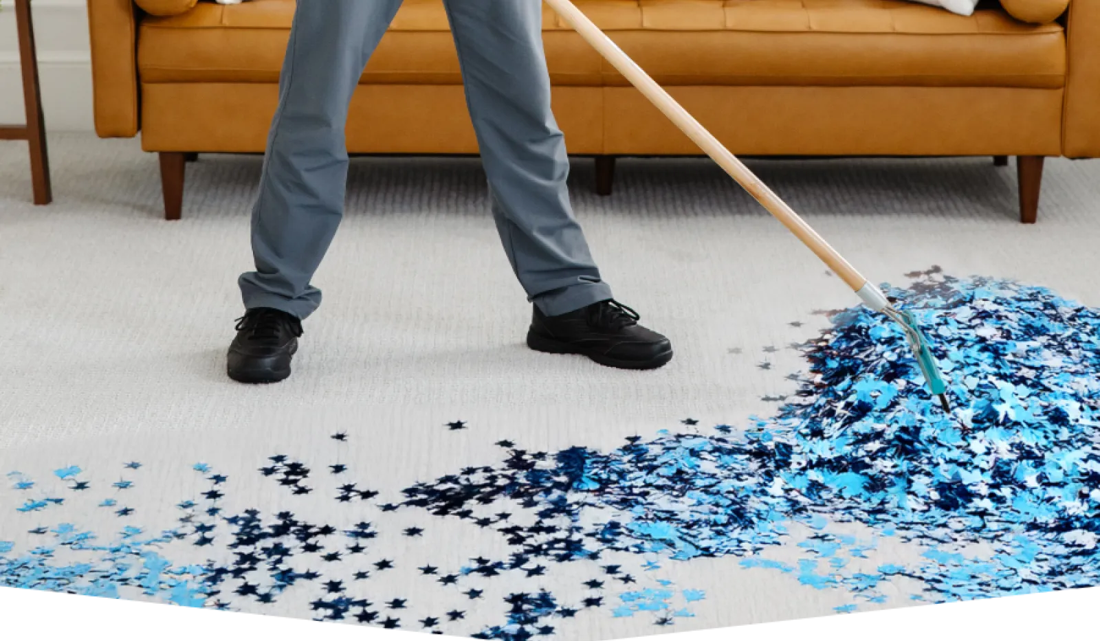 Person using a broom to sweep blue and black confetti stars on a light carpet near a brown sofa.