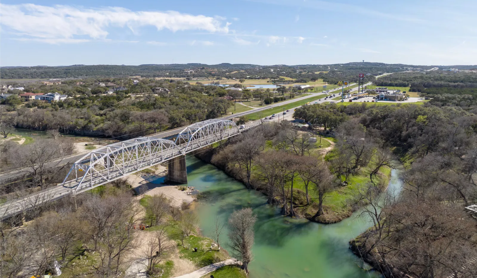 Aerial view of a white steel bridge crossing a green river surrounded by trees and roads under a blue sky.