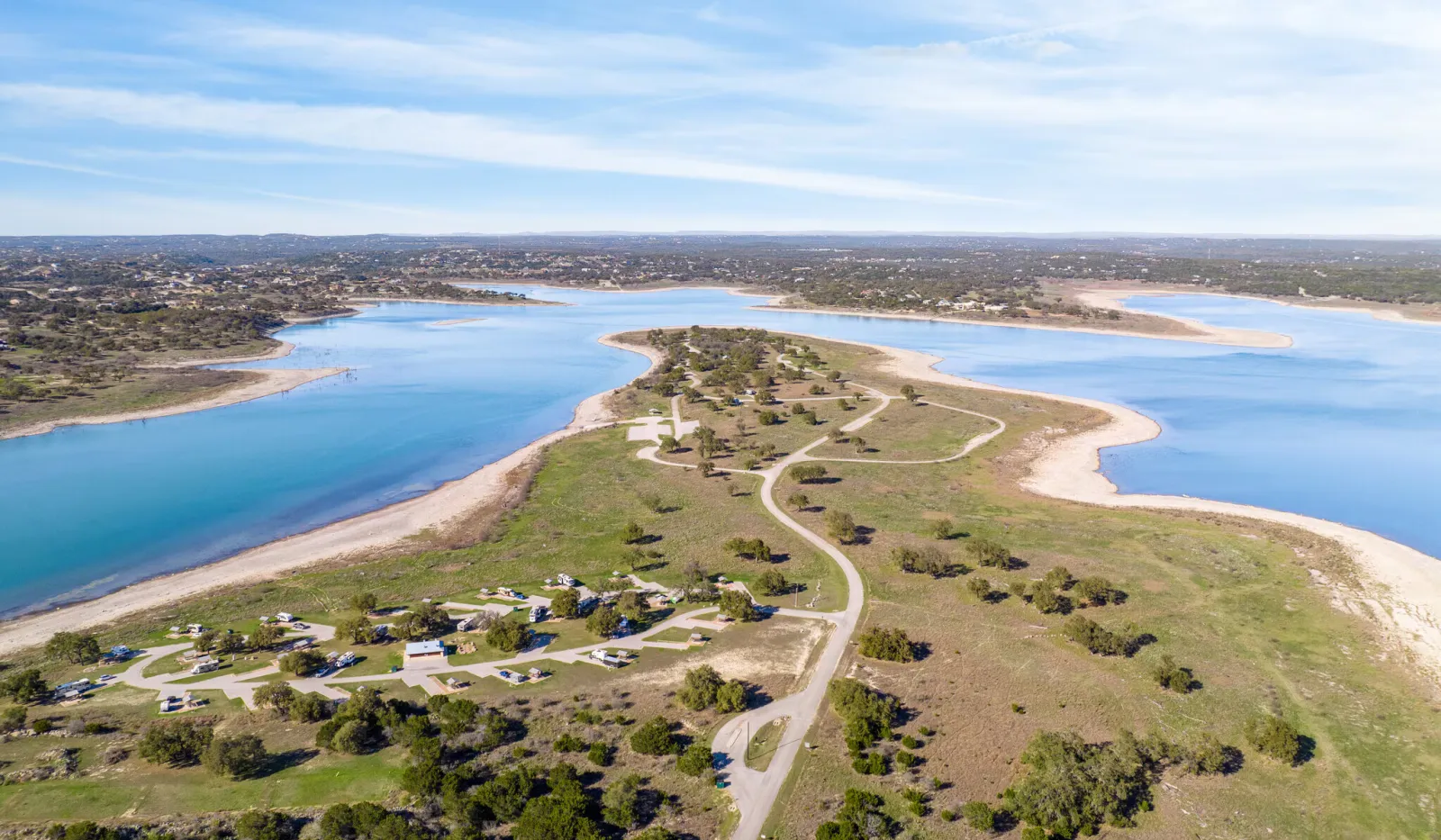 Aerial view of a winding road and green landscape surrounded by a large blue lake under a partly cloudy sky.