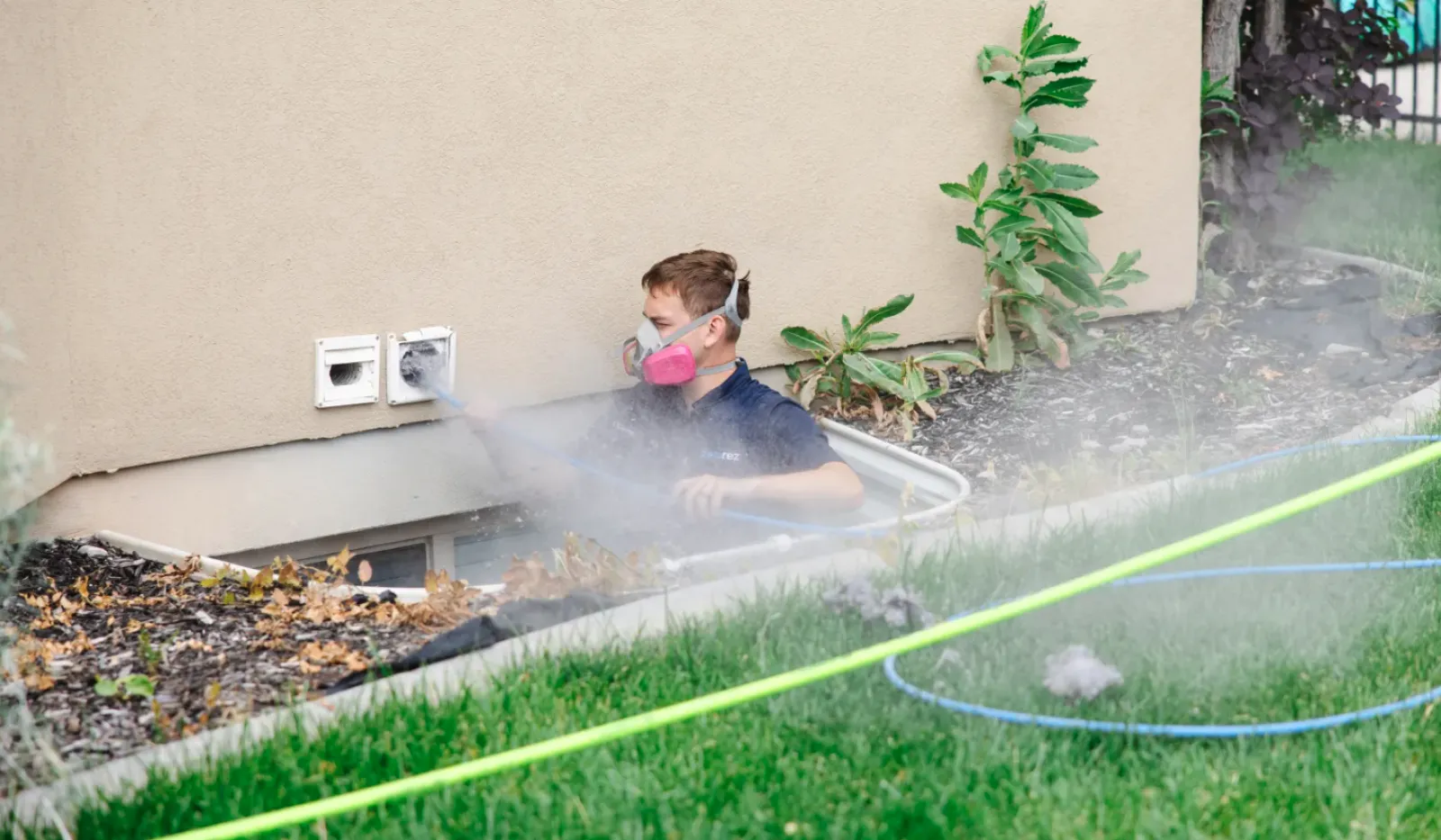 Technician wearing a mask sprays pesticide near house foundation for pest control treatment outdoors.
