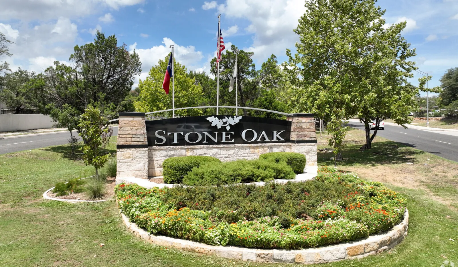 Stone Oak neighborhood entrance sign surrounded by greenery, flowers, and three flags under a blue sky.