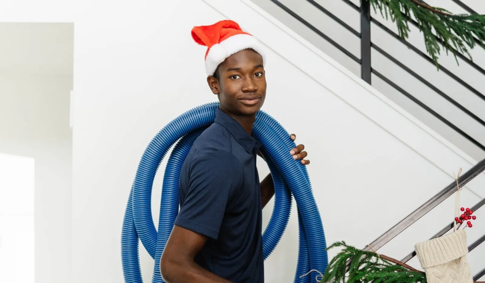Man wearing Santa hat carrying blue vacuum hose up staircase decorated with garland and white Christmas stockings.