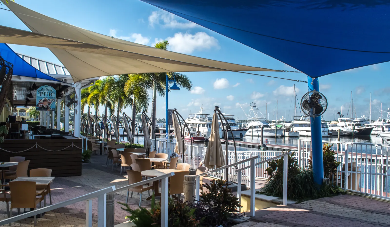 Outdoor seating area of a marina pub with shade sails, palm trees, and yachts docked in the background on a sunny day