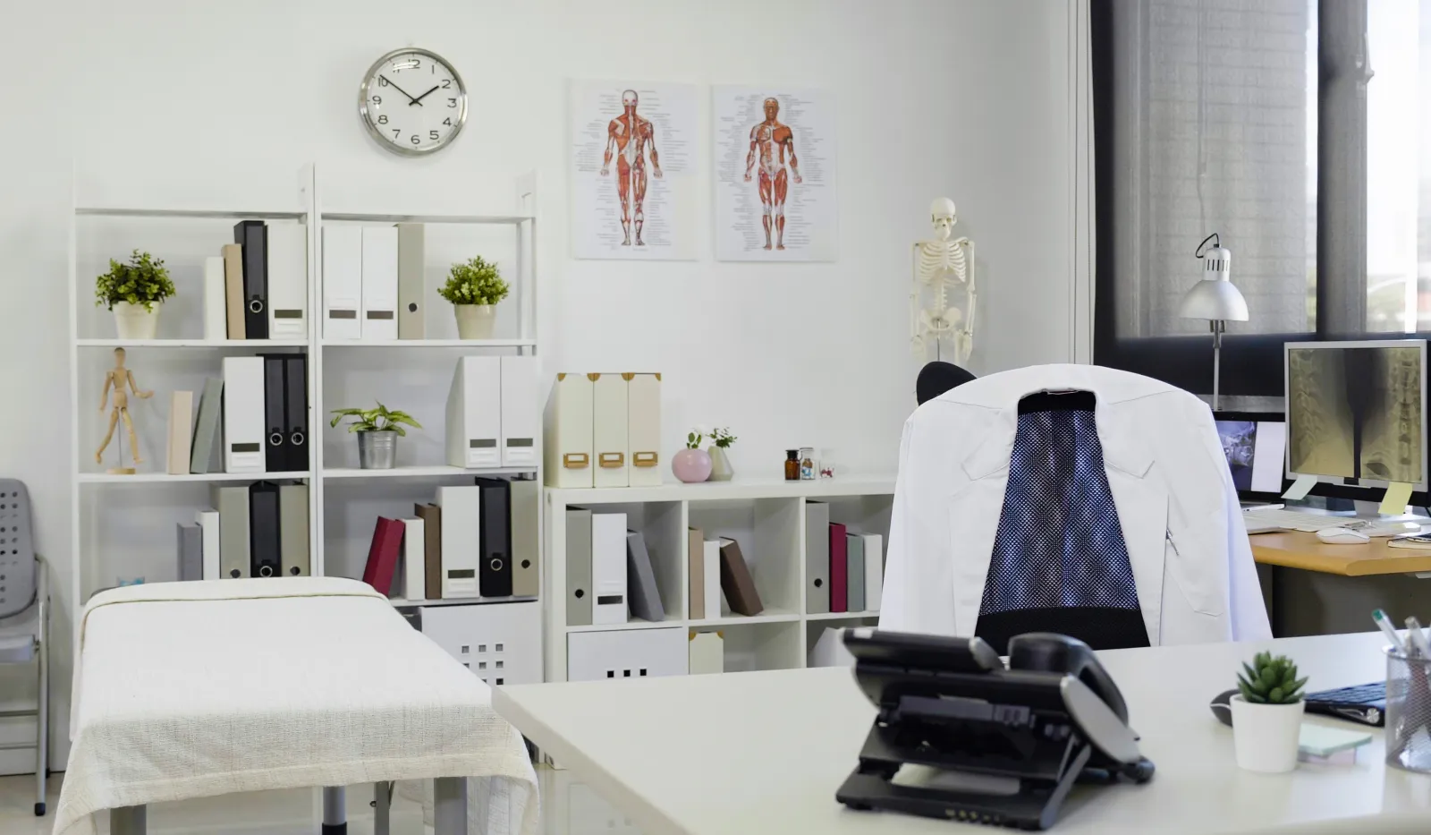 Bright medical office with an examination table, desk, computer, anatomical charts, and organized shelves.