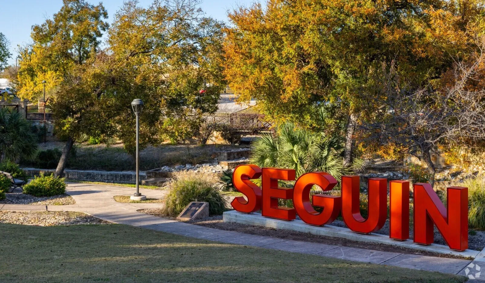 Large red SEGUIN sign in a park with autumn trees and a sunny blue sky background