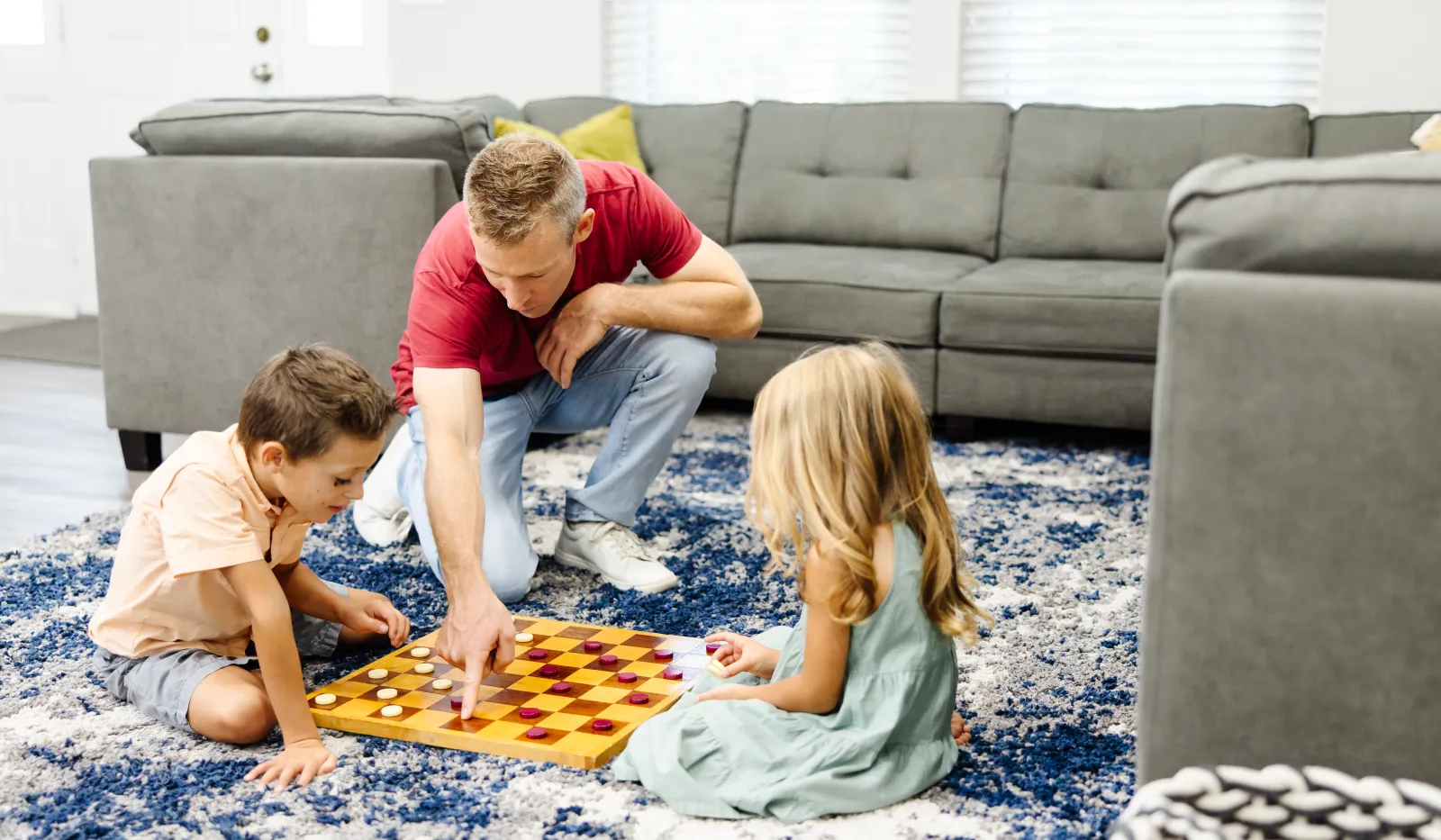 Father and two children playing checkers on a rug in a bright living room with gray sofas and cushions.