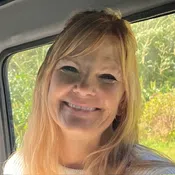 Smiling blonde woman with long hair sitting inside a vehicle with greenery visible through the window.