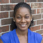 Smiling woman in a blue top with lace detail standing against a red brick wall background.