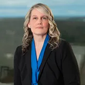 Professional woman with blonde curly hair wearing a black blazer and blue blouse in office setting.