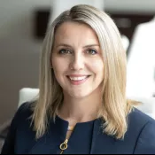 Smiling woman with blonde hair wearing a dark blazer in a bright office setting.