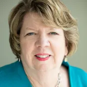 Smiling middle-aged woman with short blonde hair wearing a teal blazer and silver chain necklace.