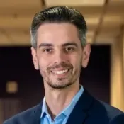 Smiling man with short dark hair wearing a blue suit jacket and light blue shirt in office setting
