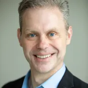 Smiling middle-aged man with blue eyes wearing a light blue shirt and black blazer.