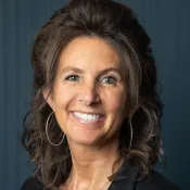 Smiling woman with curly hair wearing hoop earrings, a necklace, a black blazer, and a maroon top against dark background