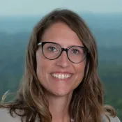 Smiling woman with glasses and wavy brown hair wearing a beige blazer, posed against a blurred natural background