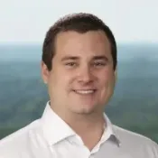 Smiling man with short brown hair wearing a white shirt, outdoors with blurred green background.