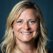 Smiling blonde woman with blue eyes wearing a cross necklace and black top against a dark background