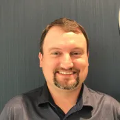Smiling man with short brown hair and goatee wearing a dark gray polo shirt against a textured dark background
