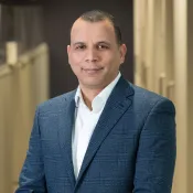 Portrait of a confident man in a blue checked blazer and white shirt standing indoors with a blurred background