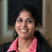 Smiling professional woman wearing a black blazer and pink patterned shirt, with dark hair and gold earrings.