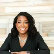 Smiling professional woman with braided hair in black blazer sitting with arms crossed indoors