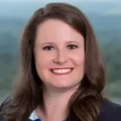 Professional woman with long brown hair wearing a blue and white floral top and dark blazer smiling outdoors.
