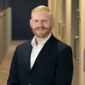 Smiling man with light hair and beard wearing a black suit and white shirt in an office corridor.
