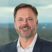 Professional middle-aged man with beard in suit jacket and white shirt smiling with blurred outdoor background.