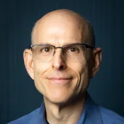 Middle-aged man with glasses and a blue shirt smiling against a dark blue background.