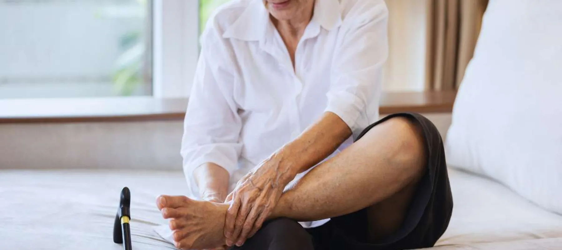 Elderly person sitting on couch holding foot in pain, with walking cane nearby in bright living room.