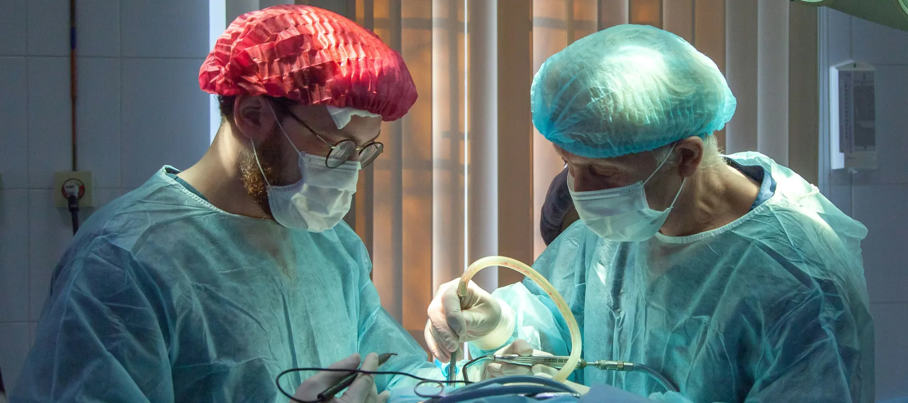 Two surgeons in scrubs and masks performing surgery under bright operating room lights.