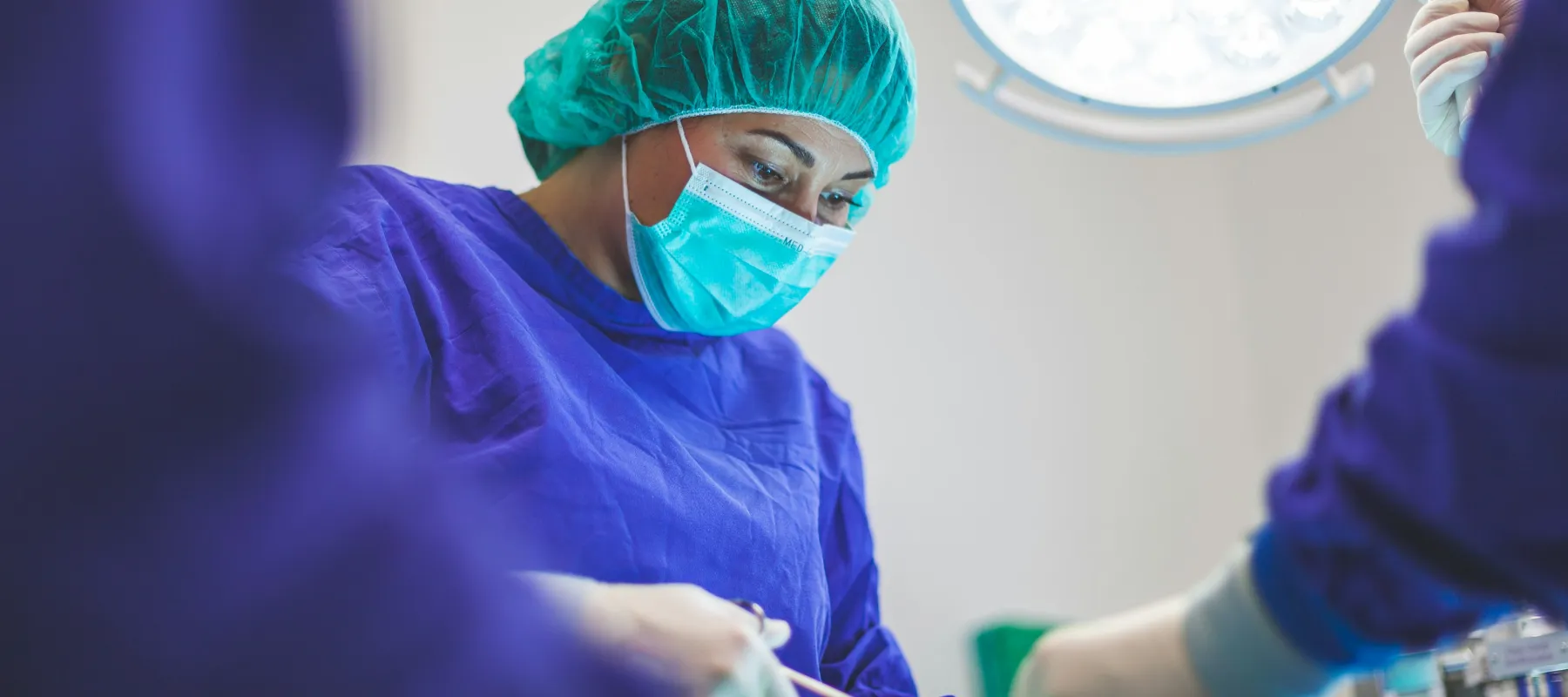 Surgeon in blue scrubs and mask performing surgery under operating room lights in a sterile environment.