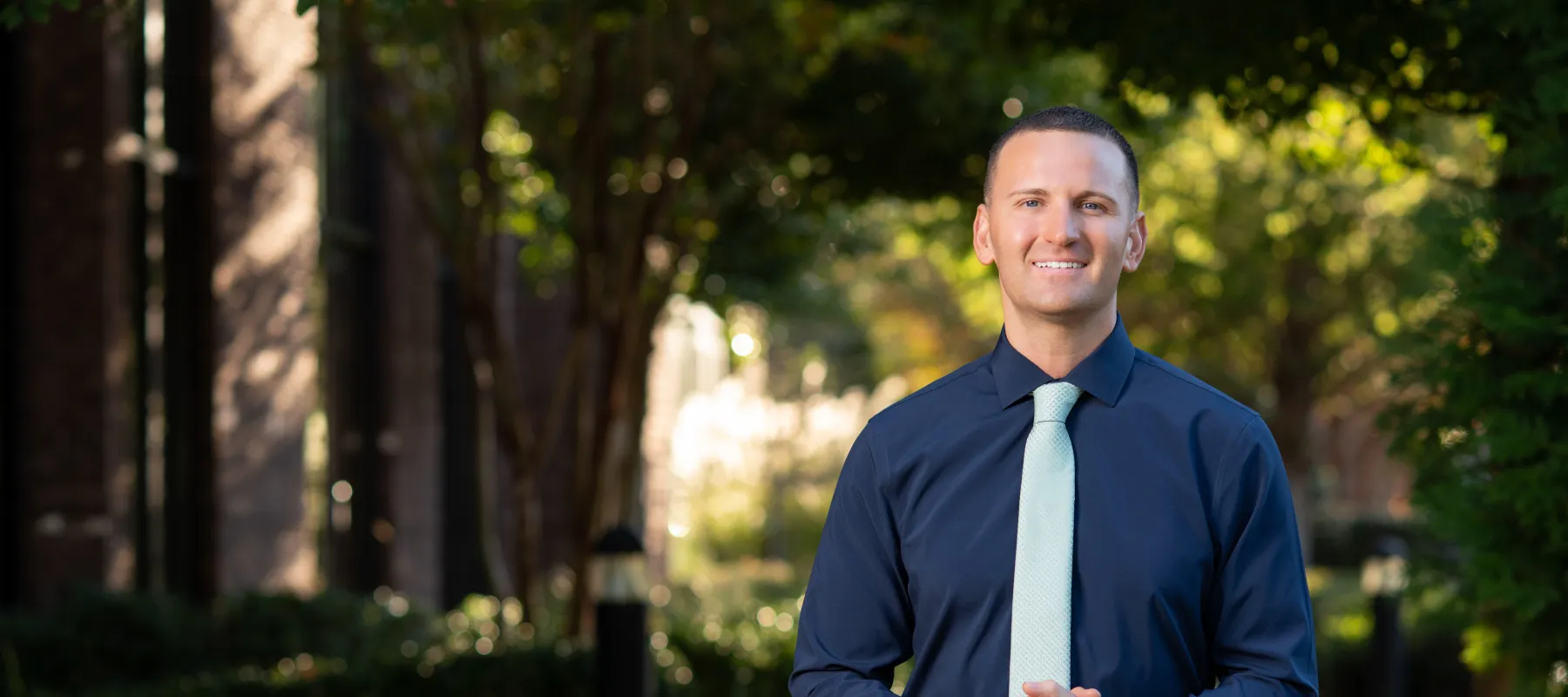 Smiling man in blue shirt with light blue tie stands outdoors on a sunny pathway surrounded by trees and bushes.