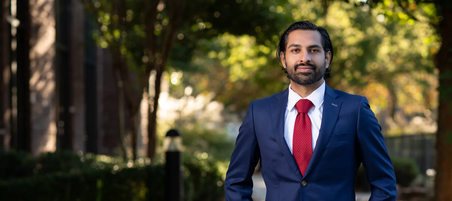 Confident man in blue suit and red tie standing outdoors with hands in pockets on a sunny day.