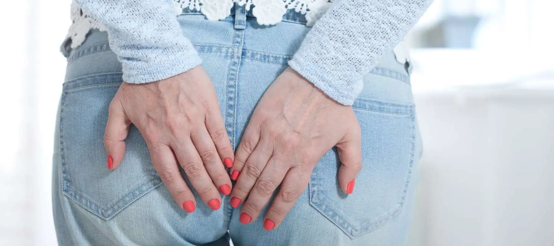 Woman with red nail polish touching her lower back while wearing blue jeans and a light sweater.
