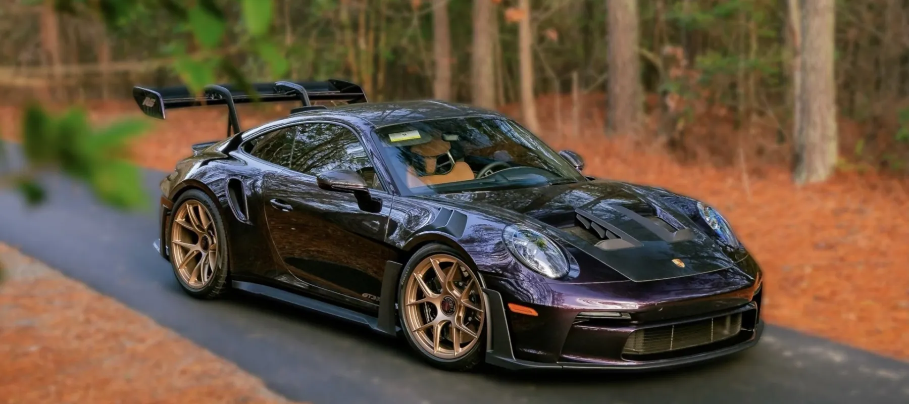 Purple Porsche GT3RS sports car with bronze wheels parked on a forest road surrounded by autumn leaves.