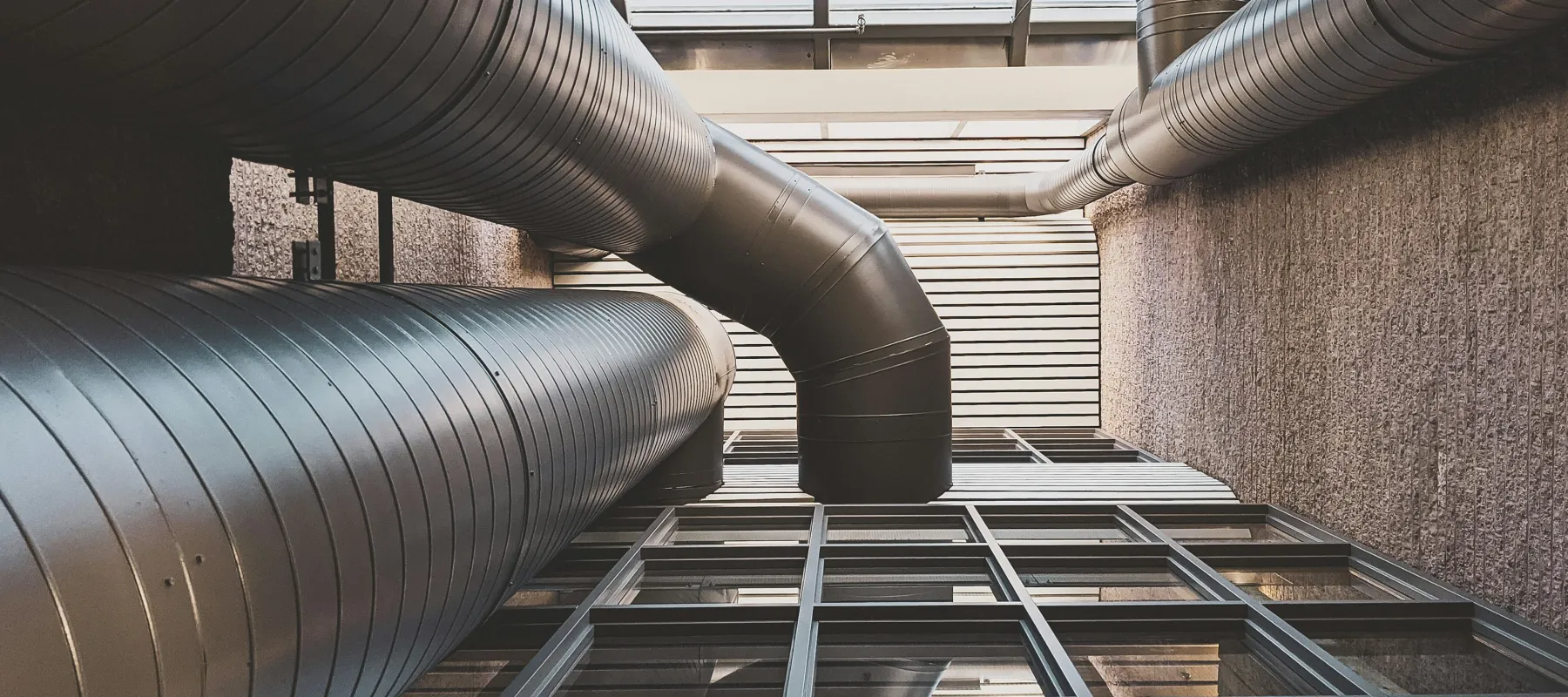 Upward view of large industrial ventilation ducts attached to a building with windows and textured walls.