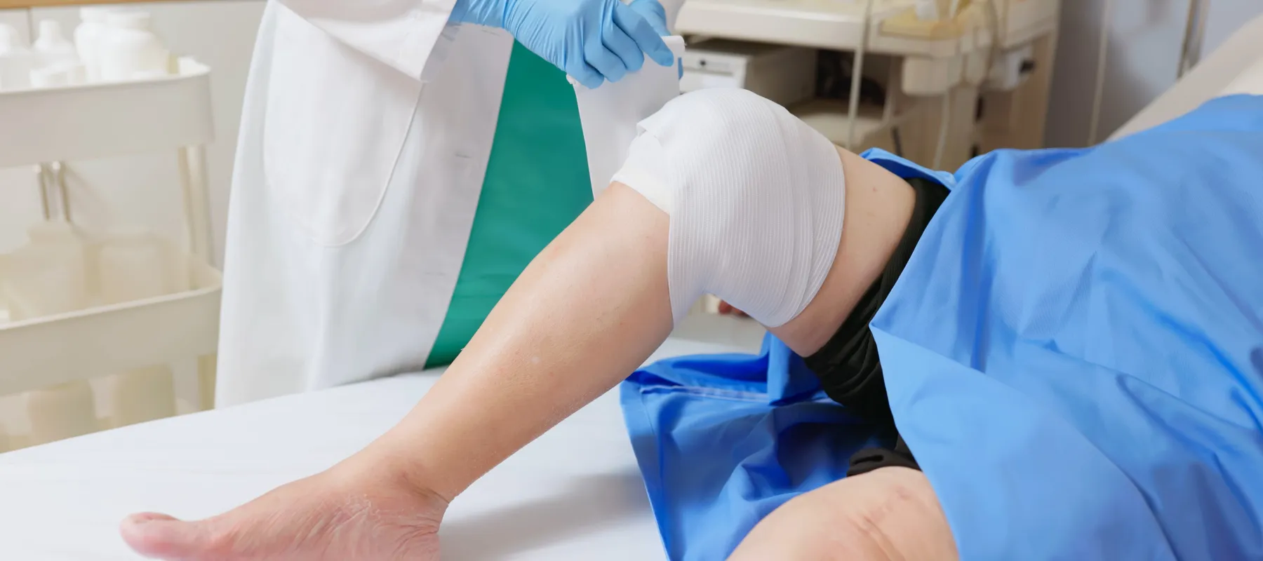 Healthcare professional adjusting bandage on patient's knee in a clinical setting with medical equipment visible.