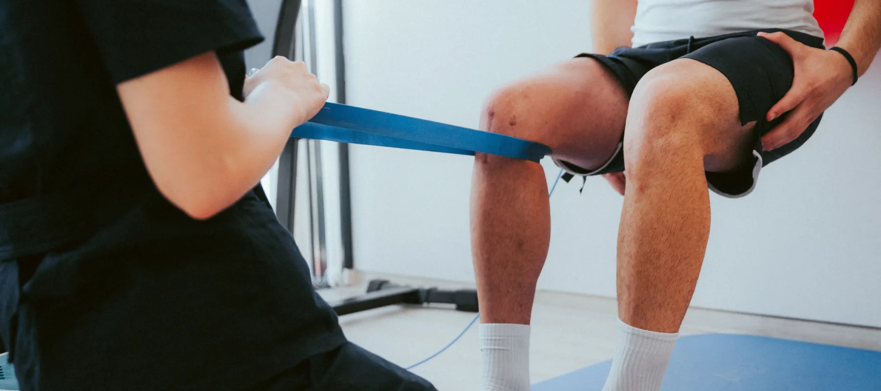 Physical therapist assisting a patient with knee resistance band exercises on a blue mat indoors.